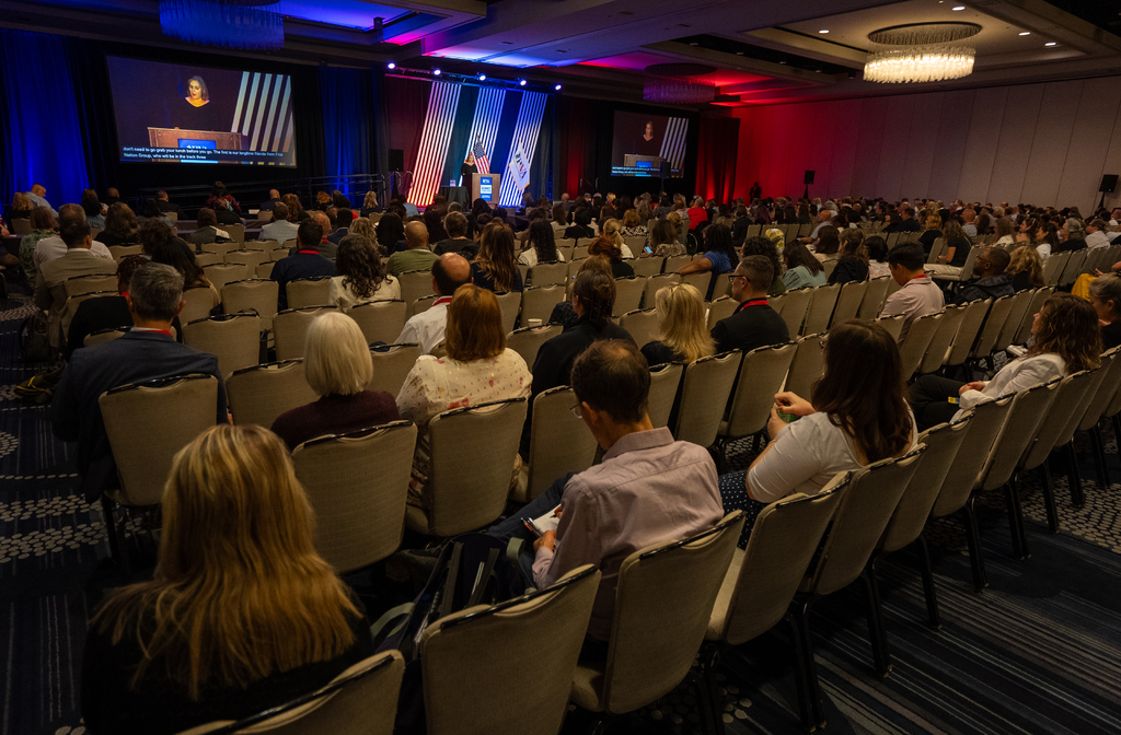 A large ballroom of attendees are seated in chairs watching a presentation being given live a large stage with two large displays on either side of the room showing content from the discussion.