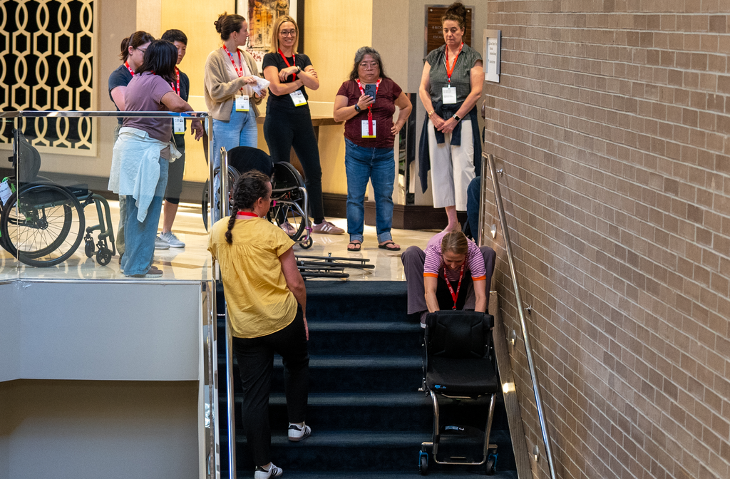 A group of attendees watch a live demonstration of a clinician showing the proper way of helping a person seated in an assisted device descend a stairwell.