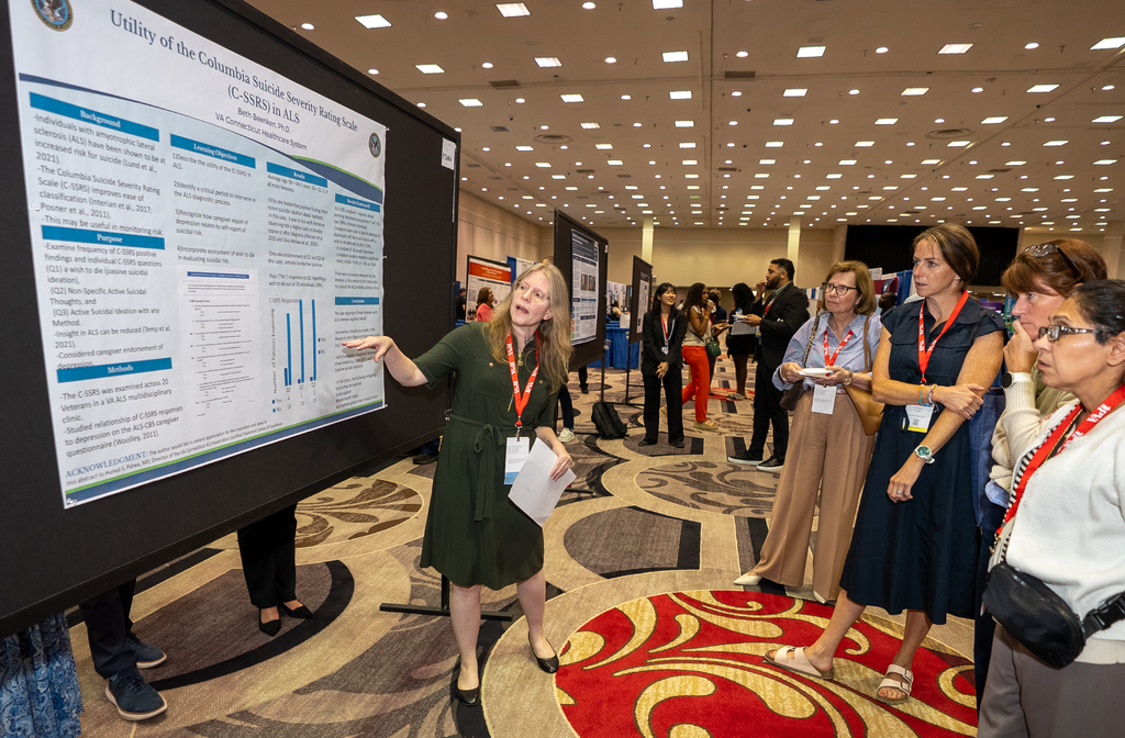 A group of attendees listen intently as a clinical researcher discusses the findings of her research presented on a large graphical posterboard.